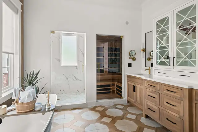 Primary bathroom with a walnut double vanity, white glass-front linen cabinet, brass sconces and a patterned tile floor