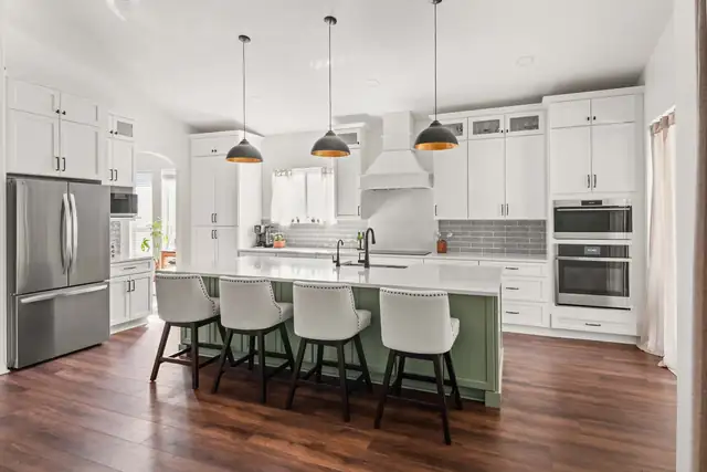 White kitchen with a sage green island, white counter stools, black dome pendants and a hood over the range