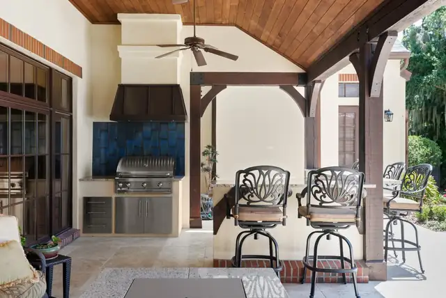 Covered outdoor kitchen with built-in grill, blue stone backsplash, black vent hood and bar stools under a wood ceiling